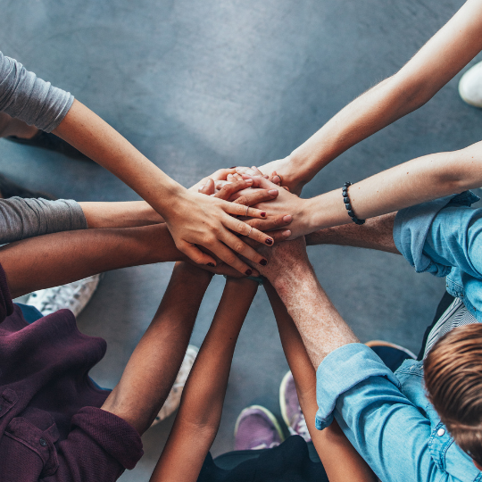 a diverse group of individuals stading in a circle, their bodies just out of camera frame but their hands allreaching into the centre of the group, stacked on top of each other in a gesture of teamwork